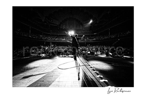 Horizontal black and white image of Steve Vai on a theatre stage with arms outstretched before a packed audience, photographed with a remote camera