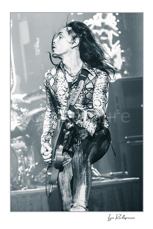 Vertical black and white image of Nuno Bettencourt performing live on stage with an electric guitar and windblown hair in dramatic motion