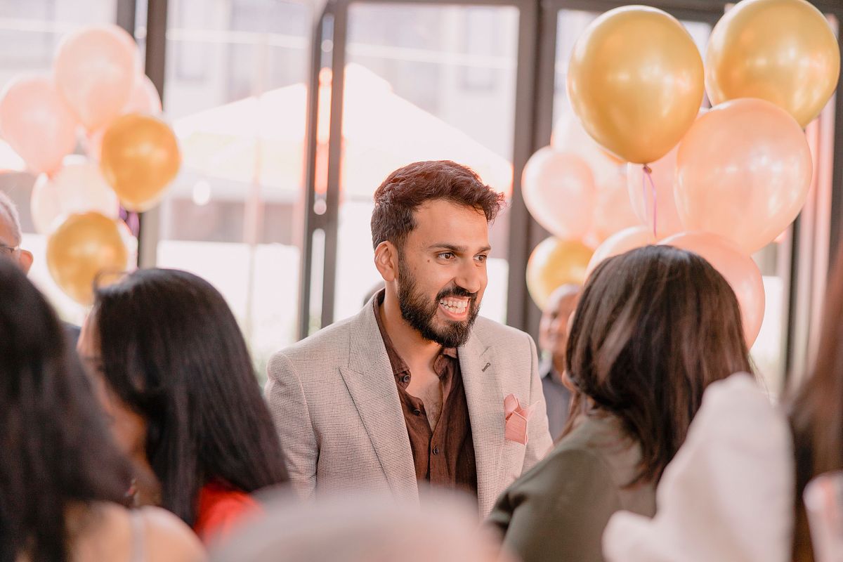 Photographer in Basel capturing a man in a light gray blazer, smiling and engaged in conversation at a lively indoor event with people and festive gold and pink balloons in the background.