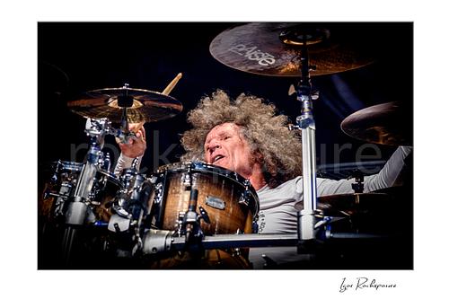 Horizontal color image of Tommy Aldridge of Whitesnake performing live on stage behind the drum kit, captured in a close-up view between cymbals and drums