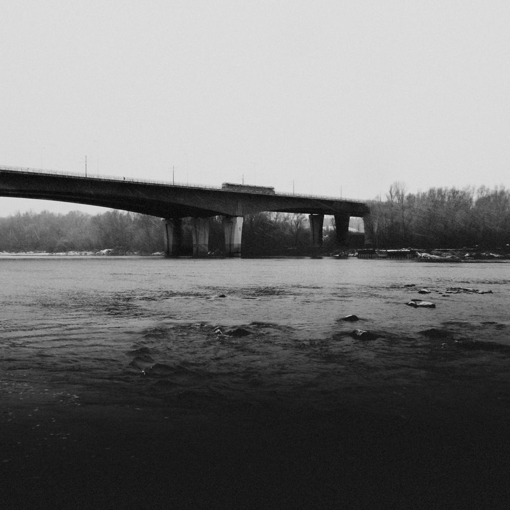 Czarno-biała fotografia autorstwa Michała Sumki ukazująca Most Północny w Warszawie na tle spokojnych wód Wisły i otaczającej przyrody. Minimalistyczna kompozycja z dominującym mostem w surowym zimowym krajobrazie.