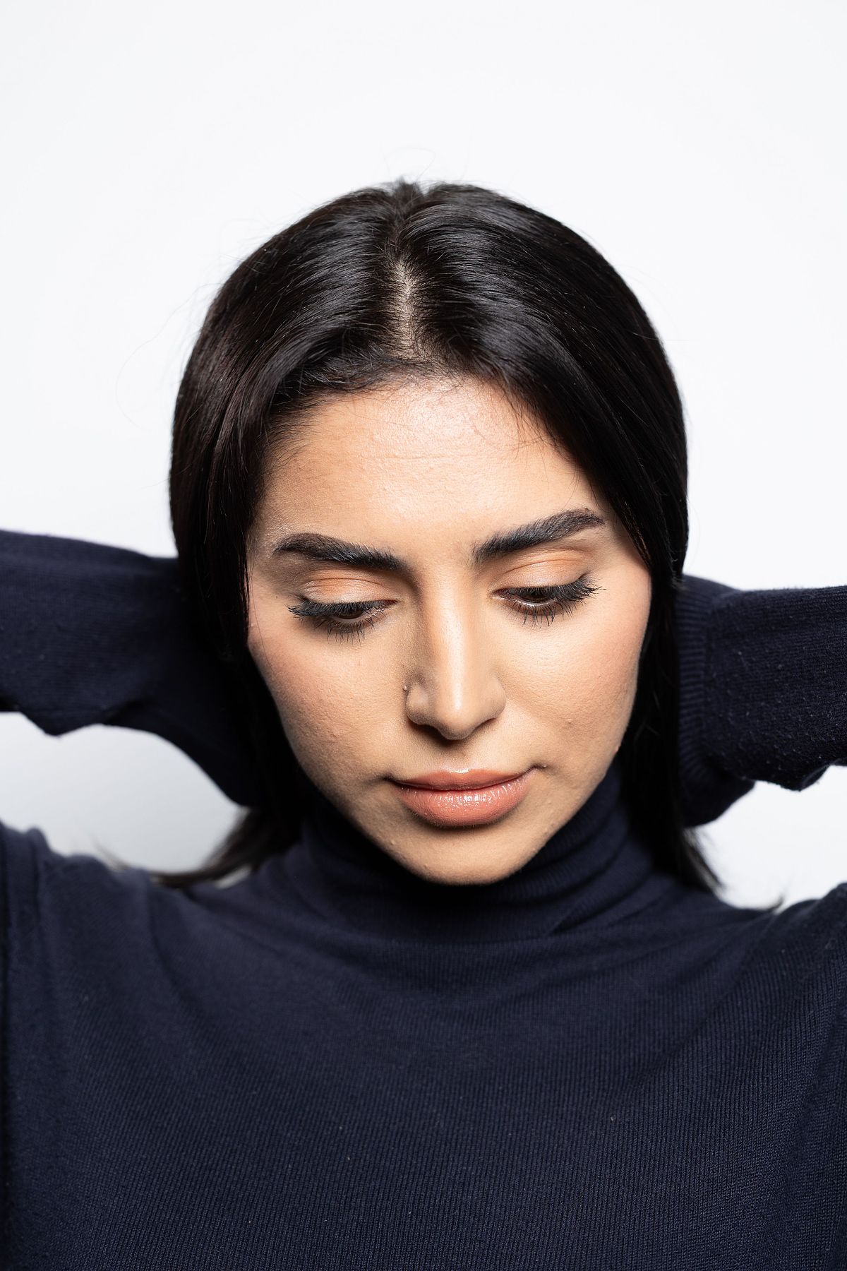 N&auml;rbild av kvinna med m&ouml;rkt h&aring;r och h&auml;nderna bakom huvudet, &ouml;gon nedslagna mot vit bakgrund