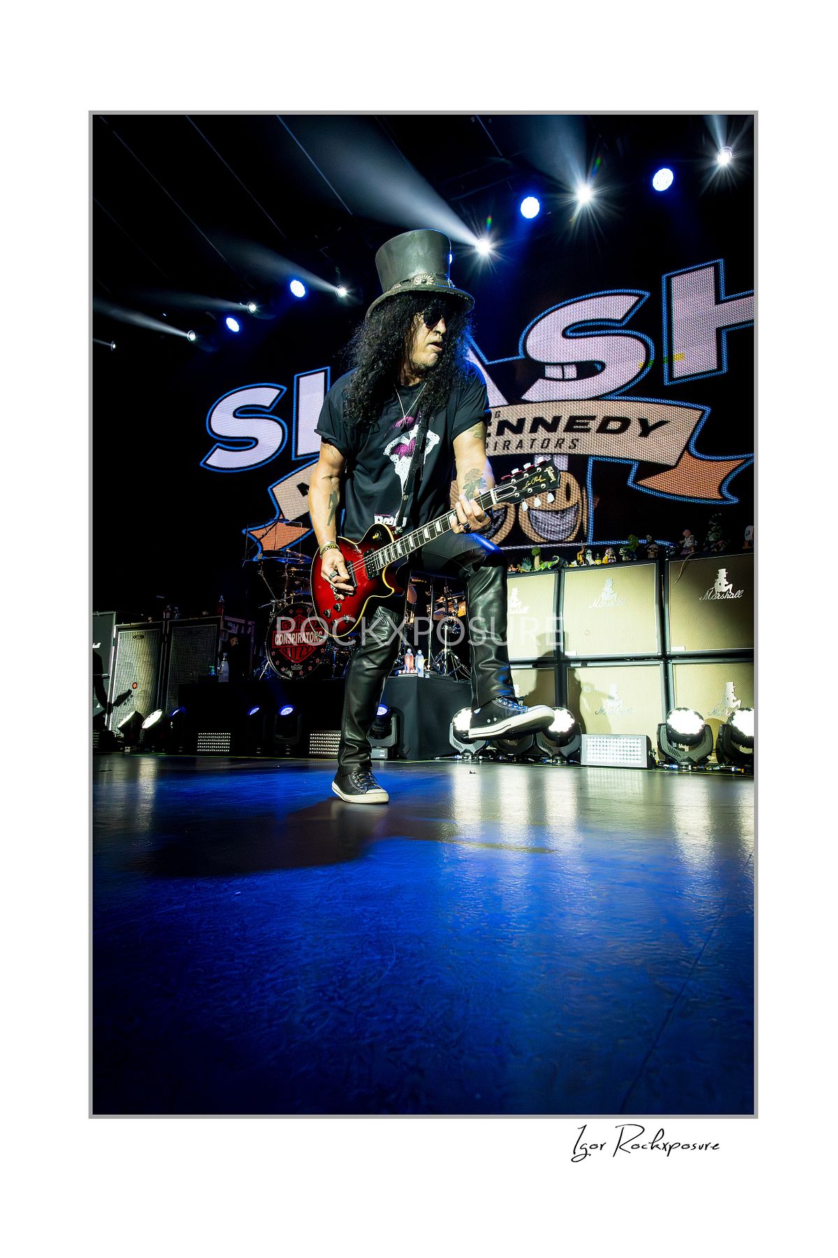 Vertical concert photography of Slash playing electric guitar in a wide stance under blue stage lighting with Slash stage backdrop