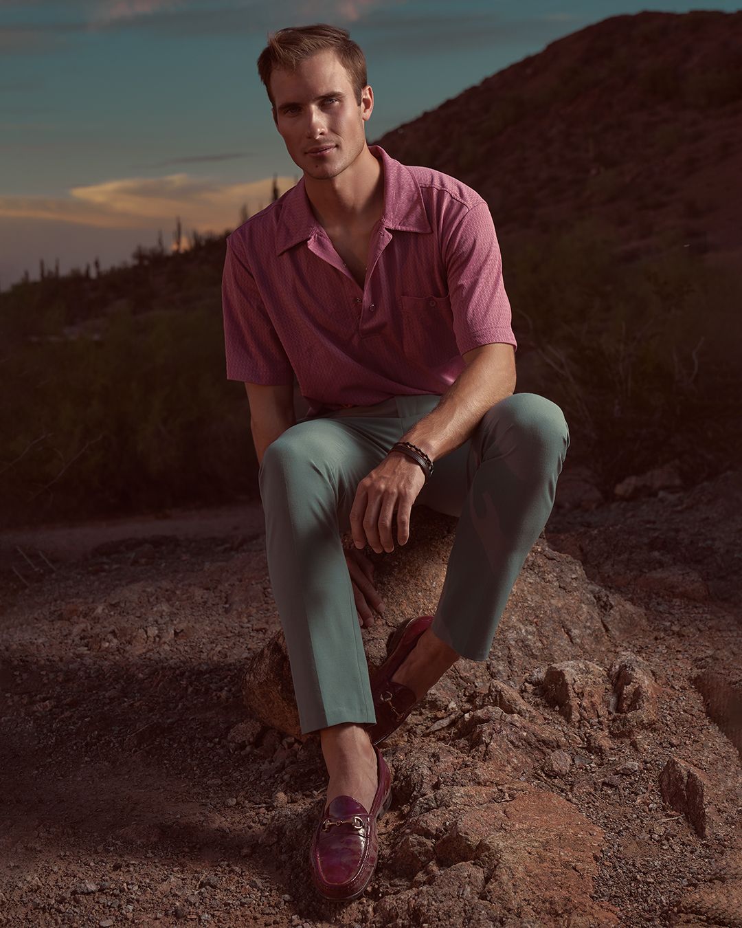 Male model sitting on rocks outdoors late in the evening in Phoenix, AZ wearing green chino pants, a pink polo shirt, and burgundy loafers