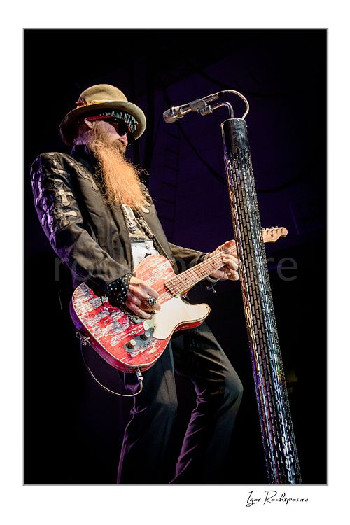 Vertical color image of Billy Gibbons of ZZ Top performing live on stage with a red Telecaster guitar, standing beside a tall textured microphone stand in his signature hat, dark glasses, and long beard
