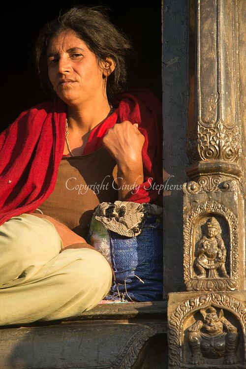 A woman with a weathered expression sits at sunset, bathed in light against a Nepalese column in a holy place for cremations in Kathmandu.