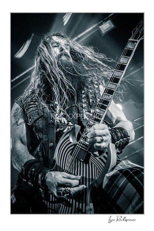 Black and white vertical close-up of Zakk Wylde playing a patterned guitar on stage, with his long hair flowing in front of his face under bright lights.