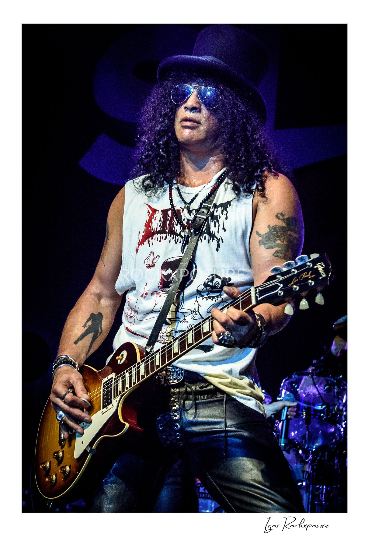 Vertical concert photography of Slash playing electric guitar wearing his signature top hat under purple and blue stage lighting