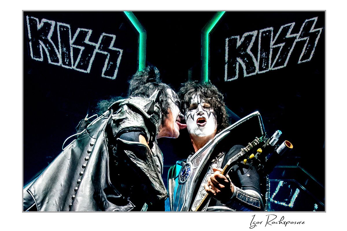 Gene Simmons of KISS leaning forward to lick Tommy Thayer’s face while Tommy plays guitar under bright blue arena stage lighting with KISS logo screens behind them