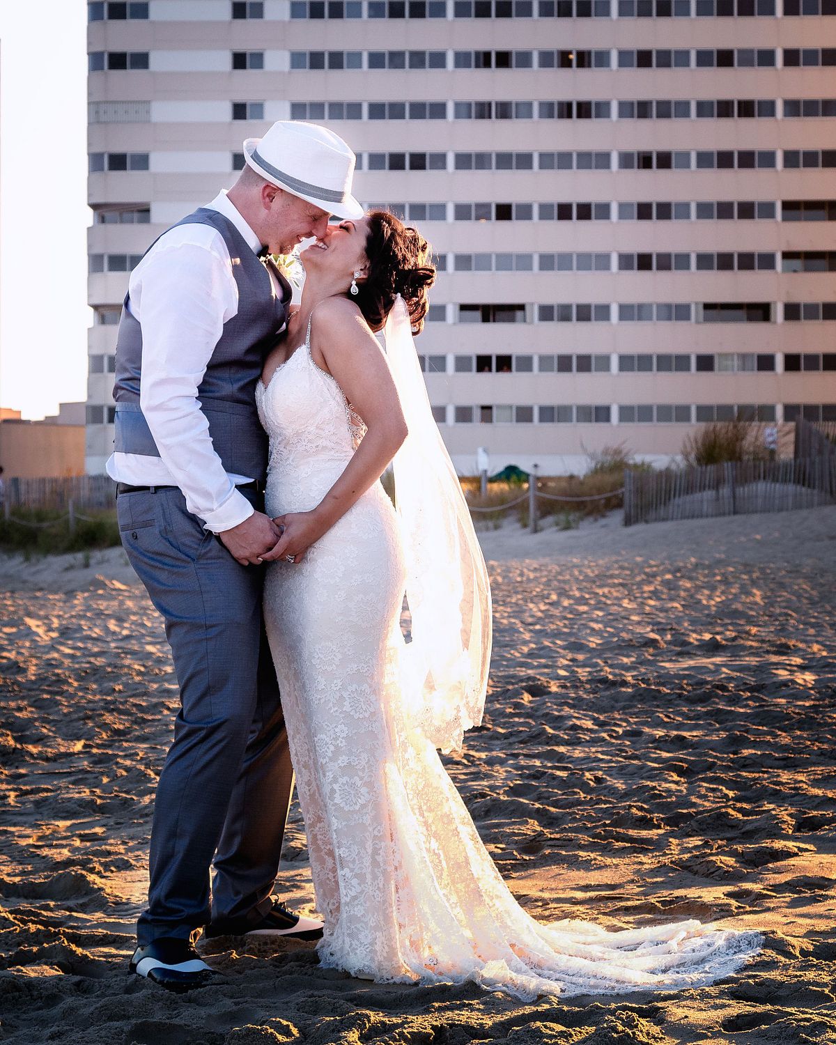 bride and groom during sunset hours on a beach in sussex county, de, the bride is wearing a long lace dress with a scalloped laced veil, the groom is wearing a hat