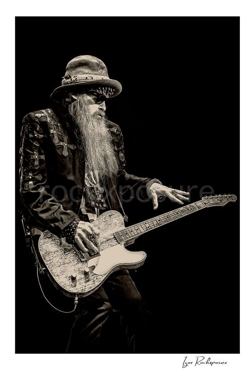 Vertical black and white image of Billy Gibbons of ZZ Top performing live on stage with a Telecaster guitar, wearing a wide-brim hat, dark glasses, and his signature long beard against a dark background