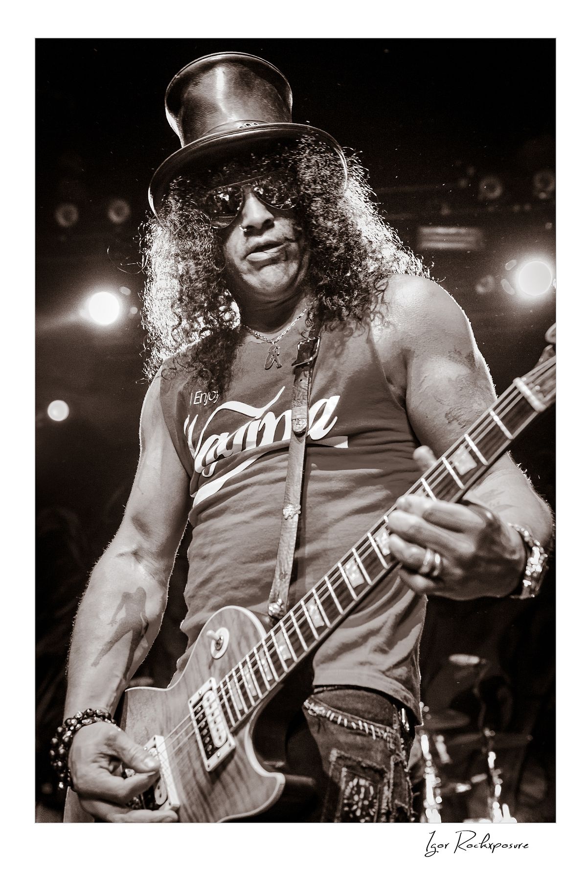 Vertical concert photography of Slash playing electric guitar wearing his signature top hat in black and white photography under stage lighting