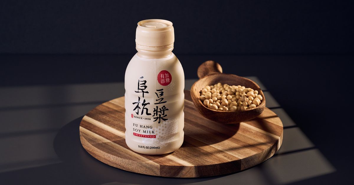 Plastic bottle of soy milk and a wooden spoon full of soy beans both sitting on a wooden dish under window light