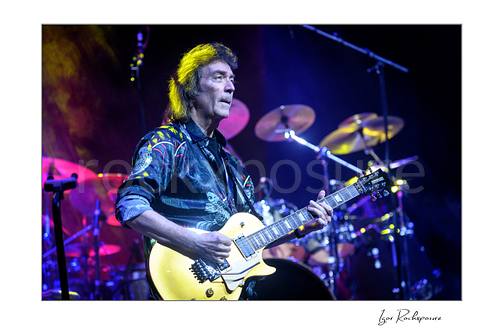 Horizontal color image of Steve Hackett performing live with electric guitar on stage with the drum kit behind him