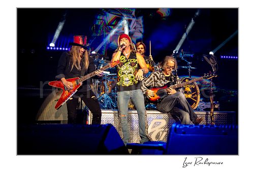 Horizontal color image of Poison performing live on stage with C.C. DeVille, Bret Michaels, Rikki Rockett and Bobby Dall under vivid blue and white concert lighting