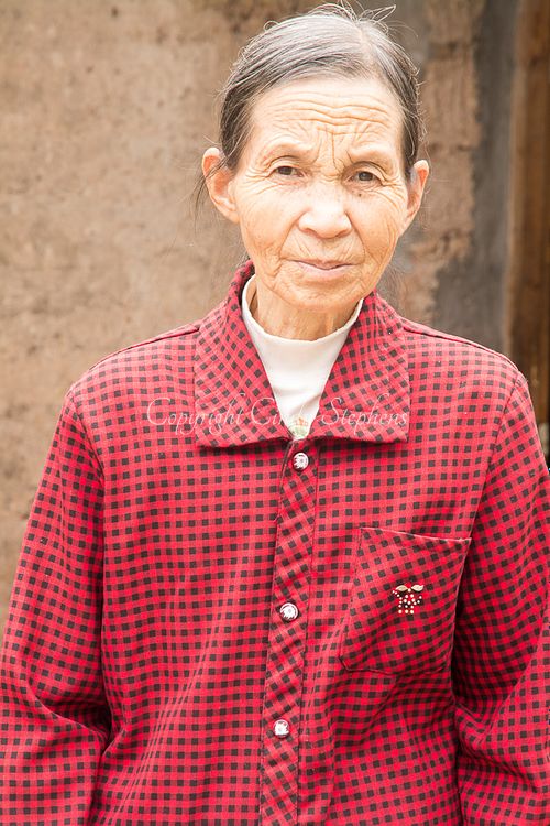 An older Chinese woman in a buttoned red shirt looks directly at the camera with a soft expression, standing outside her home in rural China.