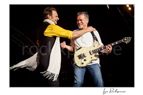 Horizontal color image of David Lee Roth and Eddie Van Halen of Van Halen sharing a live onstage moment with Eddie holding an electric guitar