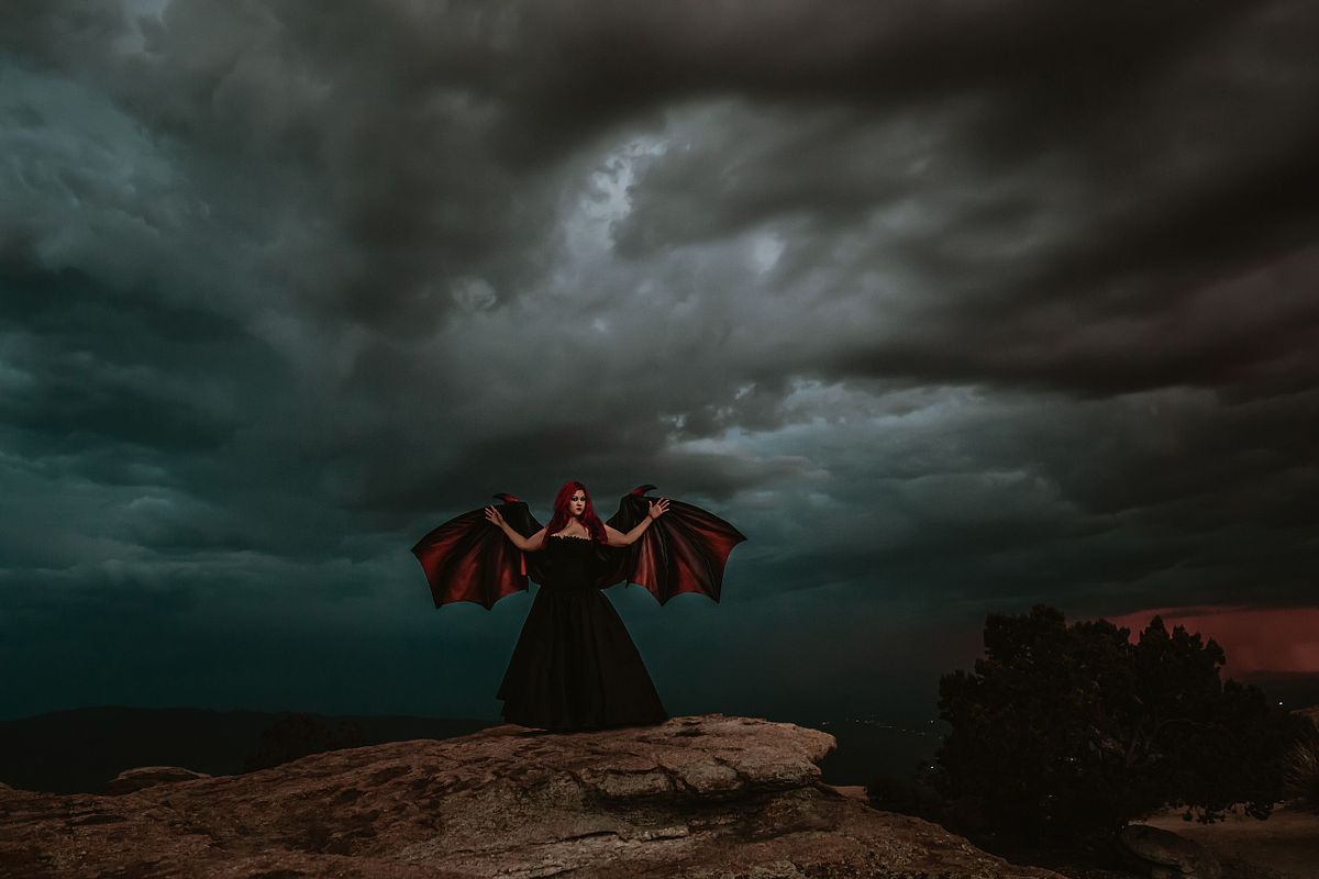 Moody and empowering boudoir portrait of a woman standing strong on a mountaintop, wearing dramatic black wings as storm clouds gather overhead — raw, fierce, and otherworldly.