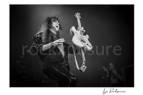 Horizontal black and white image of Yngwie Malmsteen in a dramatic live performance moment, lifting a white guitar with leopard strap under dark stage haze