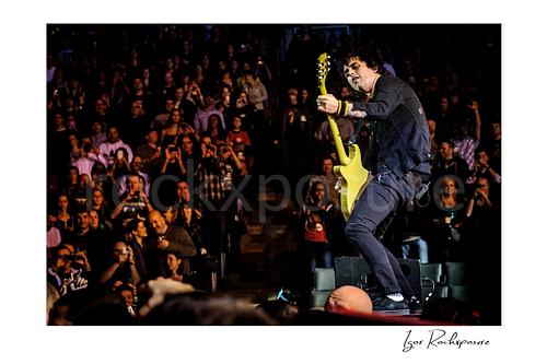 Horizontal color image of Billie Joe Armstrong of Green Day playing guitar on a stage riser with a large crowd behind him