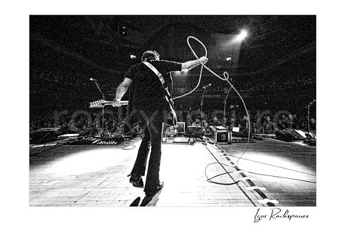 Horizontal black and white image of Steve Vai on stage from behind with guitar and a looping cable before a theatre audience