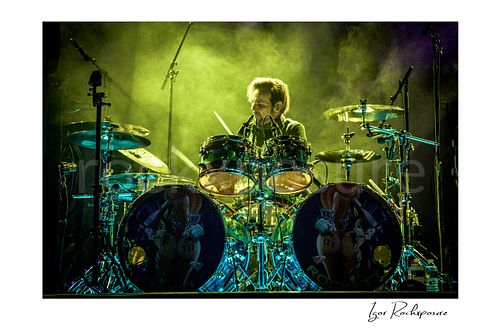Horizontal color image of Rikki Rockett performing at the drum kit during a Poison concert, surrounded by cymbals and green stage smoke