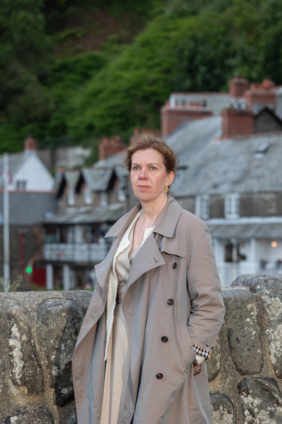 Kvinna i beige trenchcoat med kustby och tak&aring;sar i bakgrunden, editorial varum&auml;rkesportr&auml;tt.