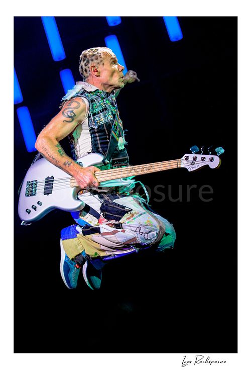 Vertical color image of Flea of Red Hot Chili Peppers suspended midair in a dramatic live performance leap, clutching his white bass against a dark stage with glowing blue LED tubes behind him