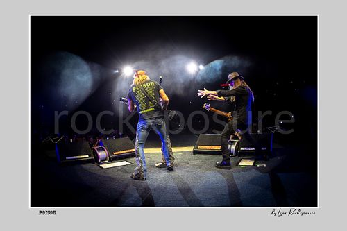 Horizontal color image of Bret Michaels and C.C. DeVille on a Poison stage, photographed with a remote camera mounted on the drum kit facing forward, with bright backlights and stage smoke