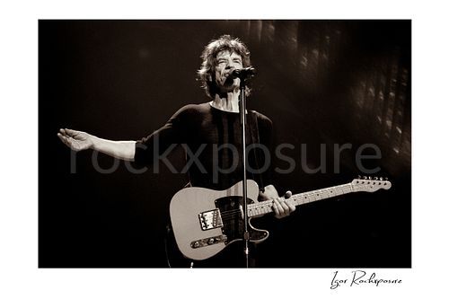 Horizontal black and white image of Mick Jagger of The Rolling Stones singing into a microphone with an electric guitar on stage