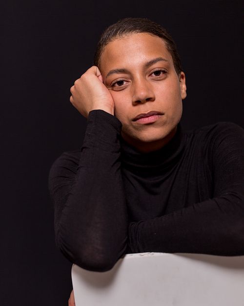 A portrait of a woman dressed in black turtle neck, sitting on a chair, with her arms placed on the white backrest.