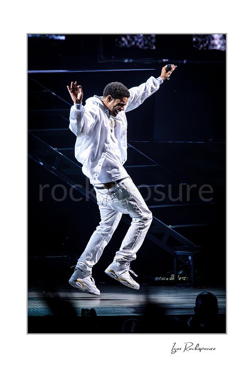 Vertical color image of Drake performing live on stage in white outfit captured mid-jump with raised arms and dramatic motion