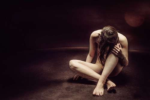 Femme nue assise au sol dans un d&eacute;cor sombre, pench&eacute;e vers l&rsquo;avant, symbolisant la culpabilit&eacute; et les cons&eacute;quences du karma
