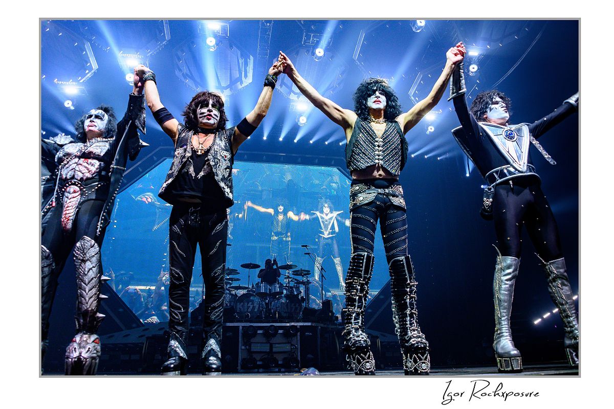 Horizontal color photograph of the full KISS band in full makeup and costumes standing with their backs to the stage and their hands raised toward the crowd as bright concert lighting fills the finale moment