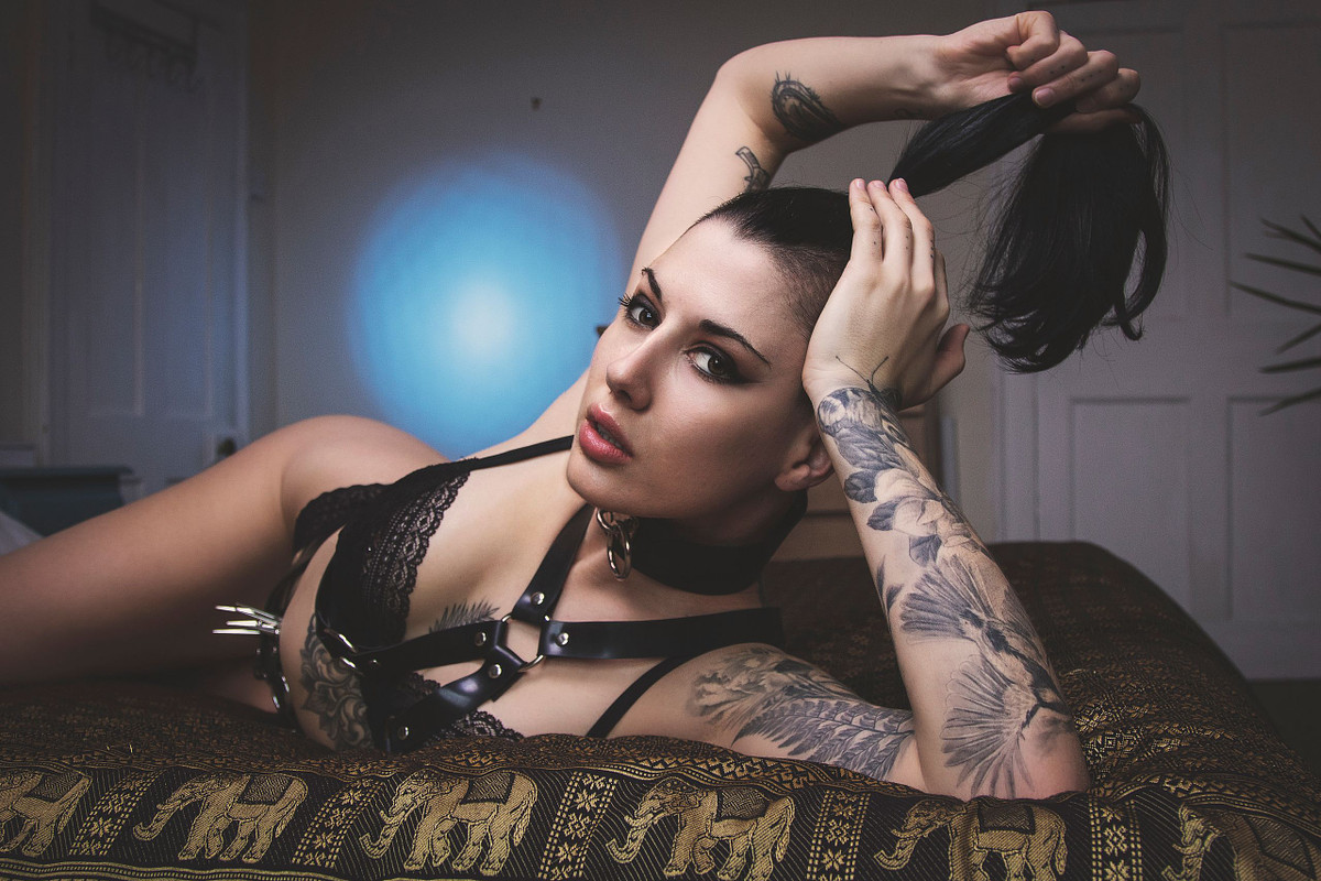 Intimate boudoir portrait of a tattooed brunette model lying on a bed with gold patterned bedding, captured by Carl Hewlett for Obsidian Noir. She is wearing black lace lingerie and a strappy leather chest harness, looking directly at the camera while holding her hair up. A circular blue light is projected on the wall behind her.