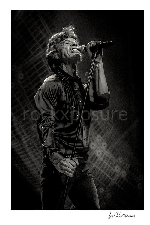 Vertical black and white image of Mick Jagger of The Rolling Stones singing into a microphone in a dramatic stage portrait