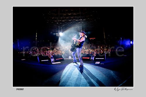 Horizontal color image of Bret Michaels on a Poison stage, photographed with a remote camera mounted on the drum kit facing forward, with bright backlight, long shadows and a packed crowd