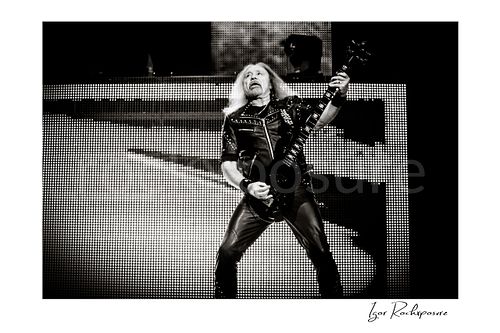 Horizontal black and white image of Glenn Tipton of Judas Priest performing live on stage, playing electric guitar in a wide stance against a bright LED screen backdrop