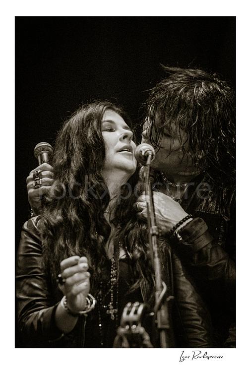 Vertical black and white image of Tom Keifer and Savannah sharing the microphone in a close live performance moment, framed tightly together at center stage