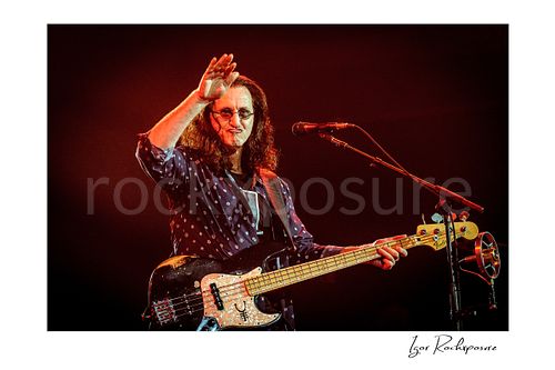 Horizontal color image of Geddy Lee performing with his bass guitar during a Rush concert, raising his hand beside a microphone under warm red stage light