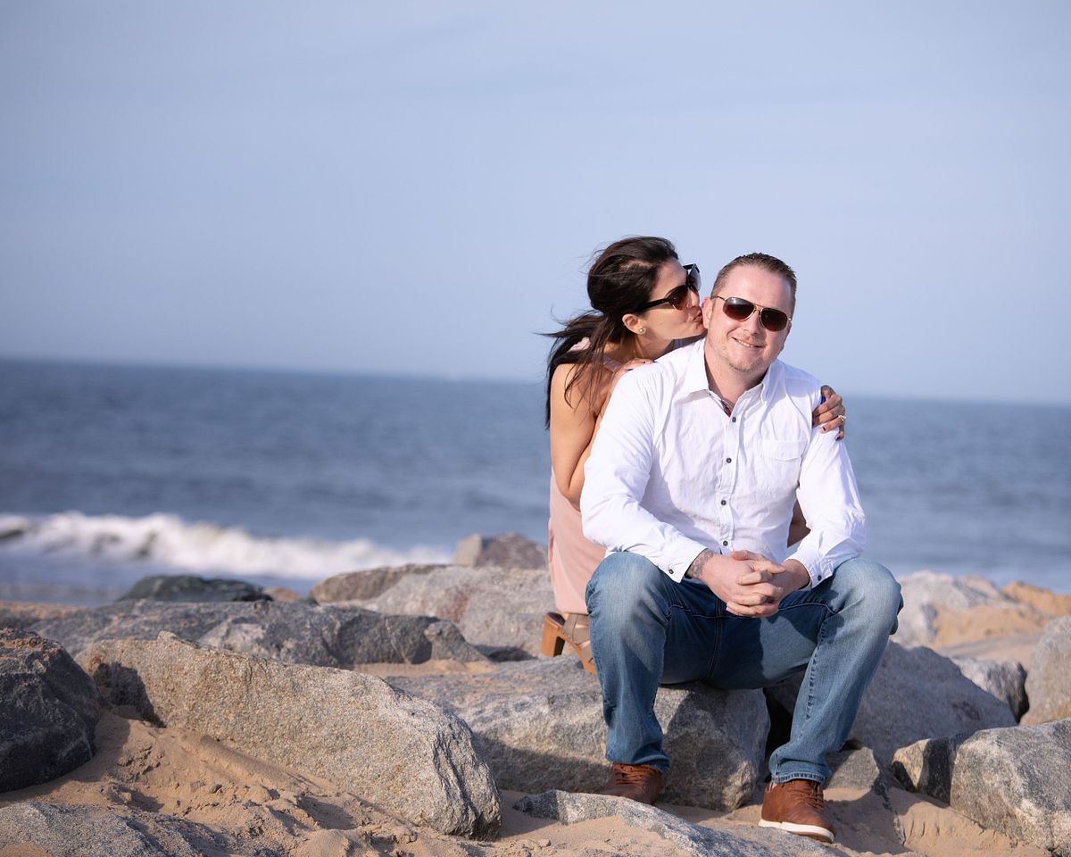 couple on the beach at cape henlopen, they are sitting on a jetty, the woman is behind the man, and kissing him on the cheek. they both are wearing sunglasses