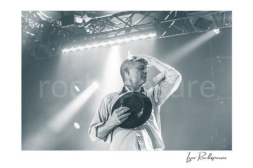 Horizontal black and white image of Gord Downie of The Tragically Hip in an emotional live performance moment, holding his hat to his chest while pressing a white cloth to his forehead under soft stage light