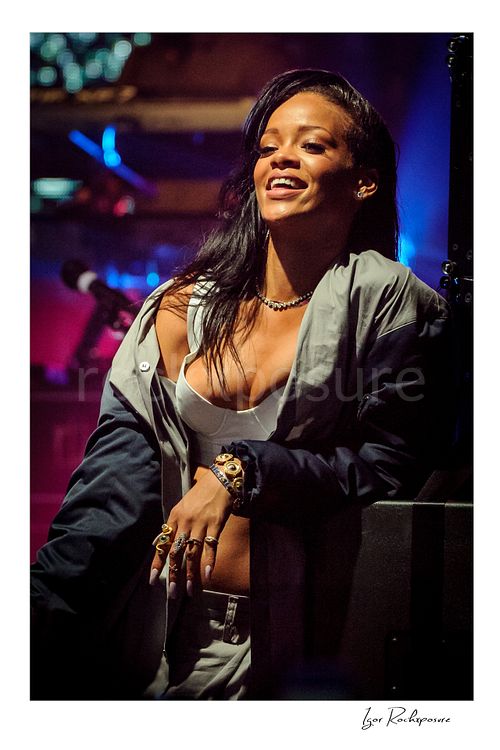 Vertical color image of Rihanna smiling on stage in a white top and draped jacket under vivid blue and magenta concert lighting