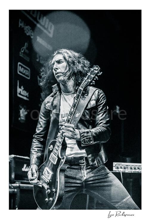 Vertical black and white image of Brent Woods playing guitar on stage wearing a leather jacket in a dramatic performance shot