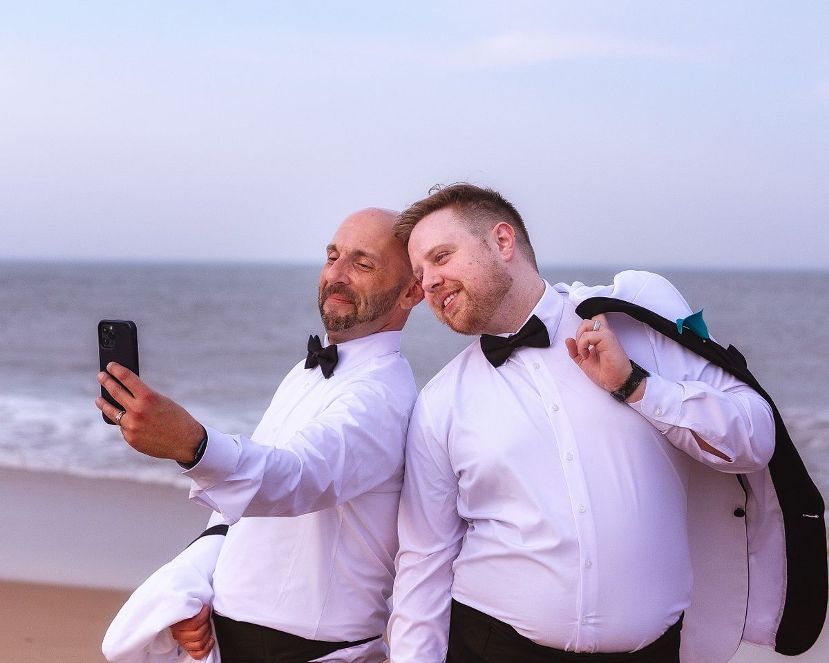 Gay couple taking a selfie at the beach, with one of the grooms having jacket draped over his shoulders