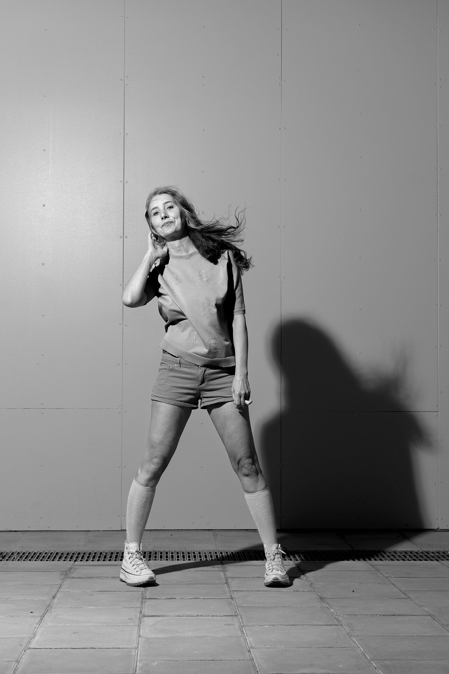 Black and white portrait of a woman in shorts standing against a plain wall with dramatic shadow, photographed in Stockholm by Mats Karlsson.