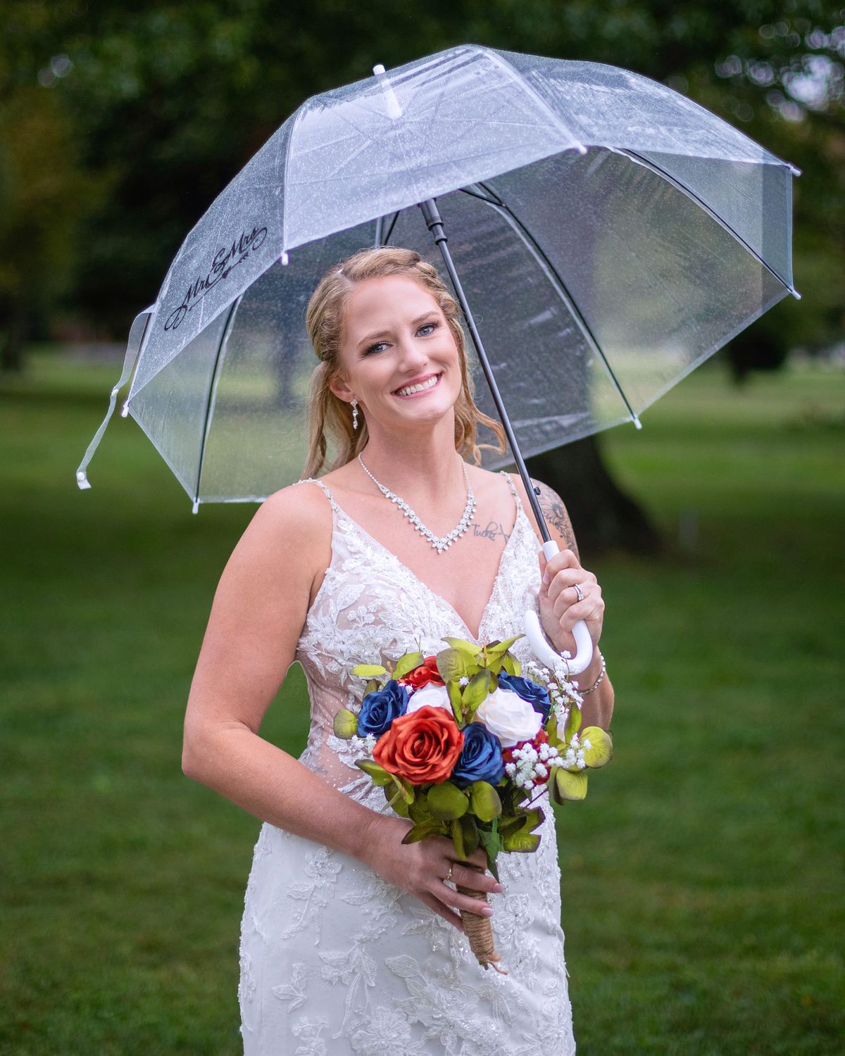 bride standing under trees holding an umbrella during a rain, she is holding her flower bouquet in her hands and is smiling