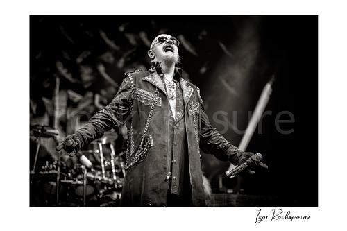 Horizontal black and white image of Rob Halford of Judas Priest performing live on stage, holding a microphone with his arms extended and the drum kit visible in the background