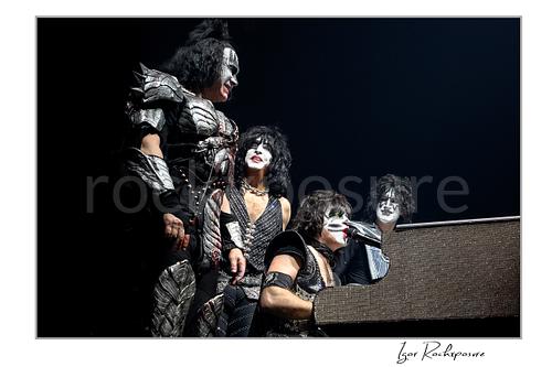 Horizontal color photograph of the full KISS band in full makeup and costumes gathered closely around Eric Singer as he plays piano and sings in a theatrical staged moment