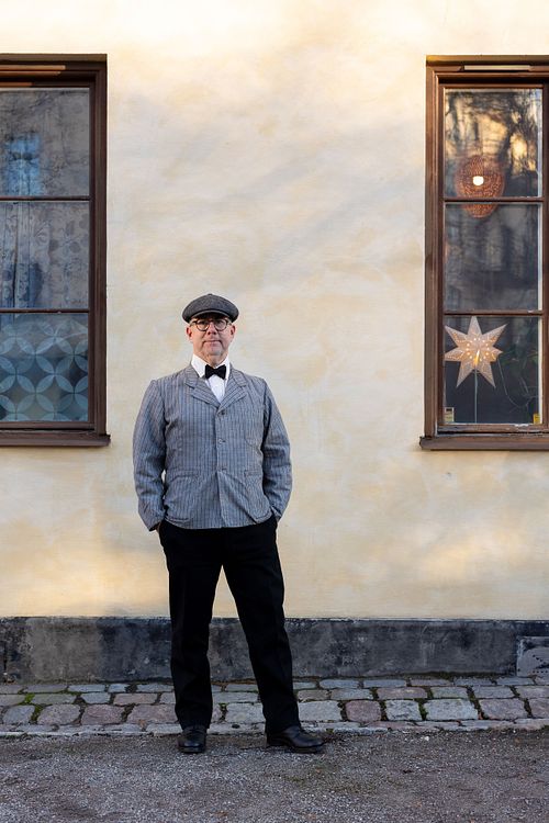 Full-body portrait of a man in a vintage-style outfit standing outside between two windows, photographed in Stockholm by Mats Karlsson.